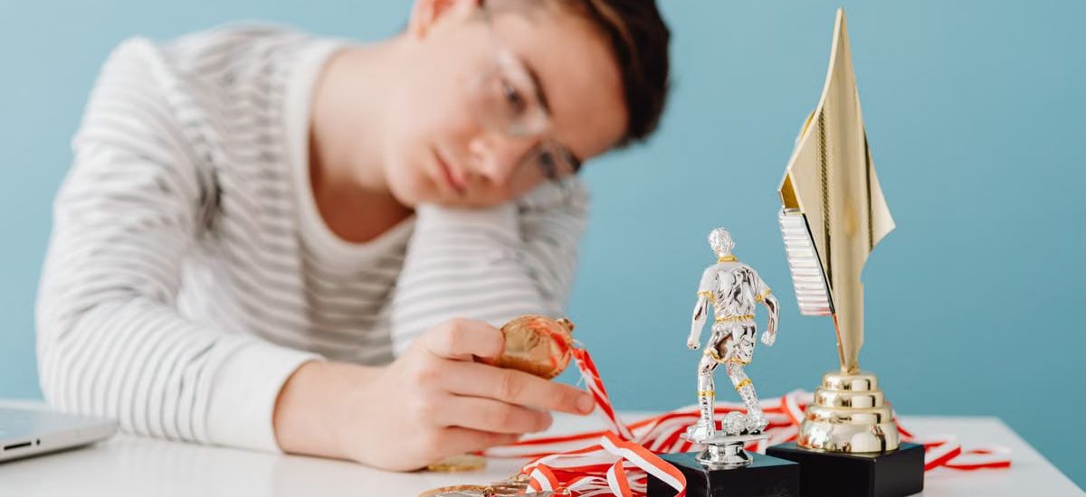 A thoughtful teenager sits by a table with various trophies and medals.